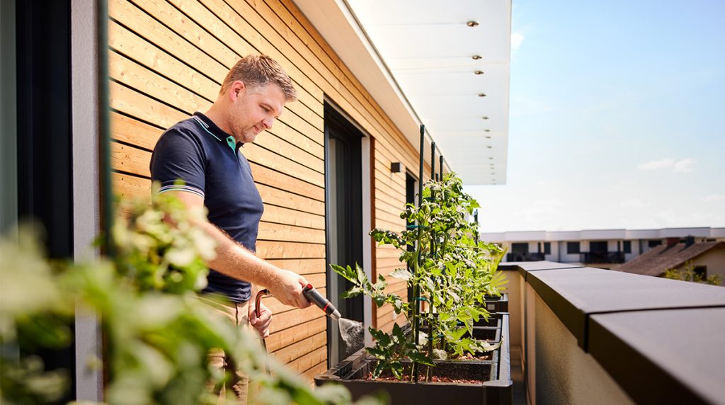 Mann lächelt, während er auf Terrasse steht und Pflanzen gießt.